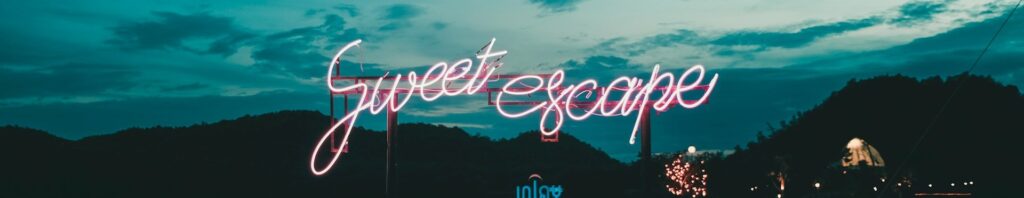 Neon lights in front of a mountain range with dusty blue sky, the lighting has the words Sweet Escape in fancy joined up pink writing.